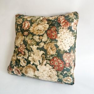 K&ouml;nnte beinhalten: Quadratisches Zierkissen mit floralem Muster. Das Kissen hat einen dunkelgr&uuml;nen Hintergrund mit gro&szlig;en Blumen in Creme-, Rot- und Beiget&ouml;nen. Die Kanten sind mit einer dunkelgr&uuml;nen Paspel versehen.