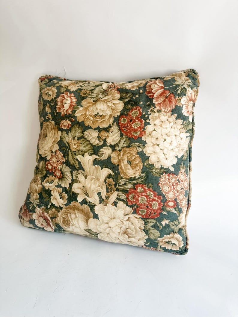 K&ouml;nnte beinhalten: Quadratisches Zierkissen mit floralem Muster. Das Kissen hat einen dunkelgr&uuml;nen Hintergrund mit gro&szlig;en Blumen in Creme-, Rot- und Beiget&ouml;nen. Die Kanten sind mit einer dunkelgr&uuml;nen Paspel versehen.