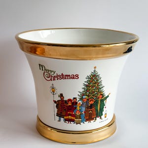 May include: White ceramic vase with gold trim at the top and base. The vase features a colorful illustration of a Christmas tree and a group of carolers, with the words "Merry Christmas" in red.