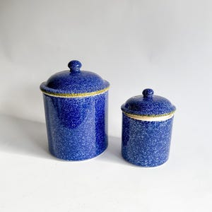 May include: Two blue ceramic canisters with lids. The canisters have a speckled pattern and a gold trim around the rim. The lids have a small knob on top. One canister is taller than the other.
