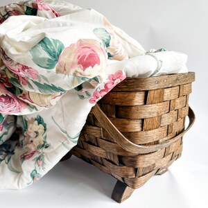 May include: A woven wooden basket with a floral patterned quilt. The quilt features pink and red roses with green leaves on a white background. The basket is brown and has a handle.