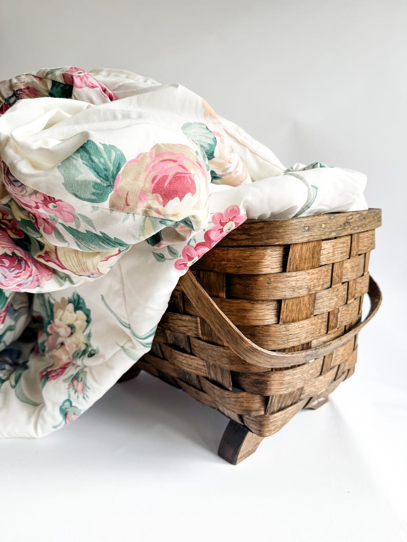 May include: A woven wooden basket with a floral patterned quilt. The quilt features pink and red roses with green leaves on a white background. The basket is brown and has a handle.
