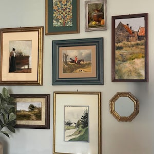 May include: A selection of framed artworks, including landscape paintings, still life, and a portrait. The frames are diverse in style and colour, with gold, brown, and green hues. The art is displayed on a pale blue wall.