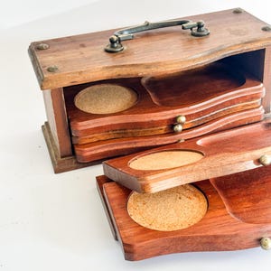 May include: A rectangular wooden storage box with a dark metal handle on top, containing multiple stacked wooden coasters. Each coaster features a circular cork inlay set into the wood. The box has a hinged design with a curved front edge.