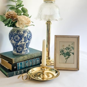 May include: A collection of vintage decor items, including a blue and white floral vase with faux flowers, a brass candlestick, a framed botanical print, and a brass lamp with a glass shade. Books are stacked in the background.