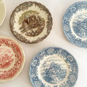 May include: A collection of four ceramic plates featuring intricate transferware patterns. One plate displays a brown landscape with a house, another a red landscape, and two plates feature blue floral borders with central rural scenes.
