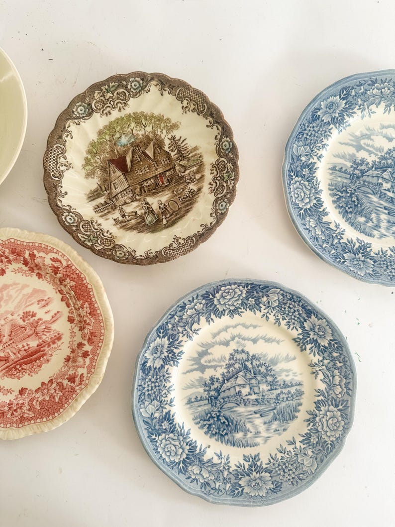 May include: A collection of four ceramic plates featuring intricate transferware patterns. One plate displays a brown landscape with a house, another a red landscape, and two plates feature blue floral borders with central rural scenes.