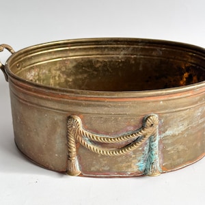 May include: An antique brass planter with a weathered patina. The oval-shaped container features decorative rope and tassel accents. Two small handles are on either side. The planter's aged appearance suggests it is vintage.