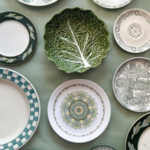 May include: A collection of ceramic plates and a bowl featuring various designs, including a green cabbage-shaped bowl, a plate with a Williamsburg Virginia illustration, a checkered rim plate, and several floral and patterned decorative plates.