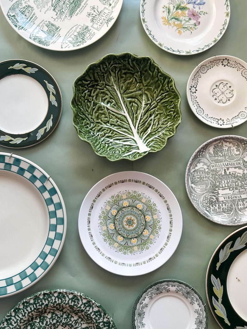 May include: A collection of ceramic plates and a bowl featuring various designs, including a green cabbage-shaped bowl, a plate with a Williamsburg Virginia illustration, a checkered rim plate, and several floral and patterned decorative plates.