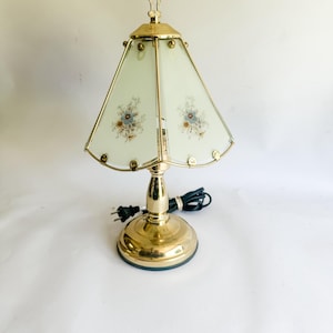 Peut inclure: Une lampe de table vintage en laiton avec un abat-jour à motifs floraux. L'abat-jour est de couleur vert clair avec des bordures dorées et présente des motifs floraux bleus et bruns. La lampe a une base ronde et une poignée décorative.