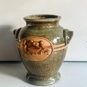 以下が含まれることがあります： 緑と茶色のセラミック花瓶。鳥と巣のレリーフが特徴です。花瓶には2つのハンドルと装飾的なバンドがあります。全体的なデザインは素朴で自然にインスパイアされています。花や装飾品に最適です。