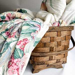 May include: A wooden basket filled with a floral patterned quilt. The quilt features pink, red, and green flowers on a white background. The basket is made of woven wood and has a handle.