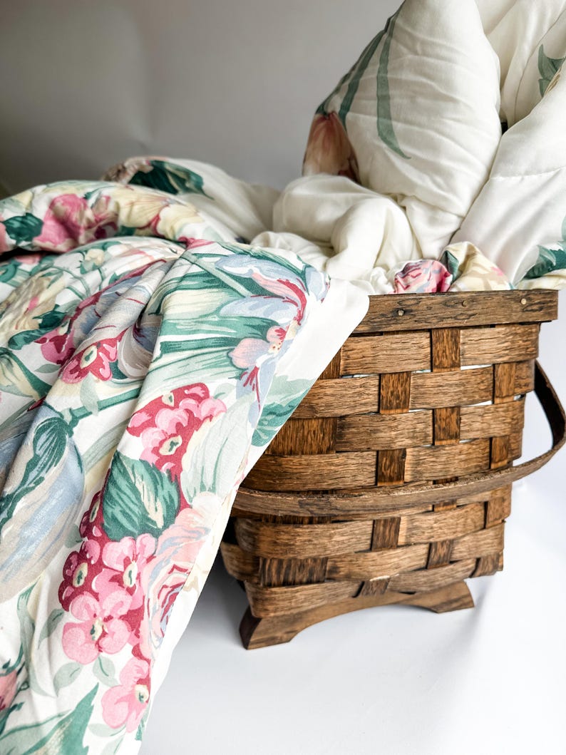 May include: A wooden basket filled with a floral patterned quilt. The quilt features pink, red, and green flowers on a white background. The basket is made of woven wood and has a handle.