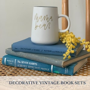 May include: A white ceramic mug with "mama bear" in gold script sits atop a stack of vintage blue books. A sprig of yellow flowers adds a pop of color. The books are on a woven surface.