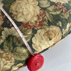 K&ouml;nnte beinhalten: Ein Kissen mit floralem Muster mit einem Ma&szlig;band, das ein Ma&szlig; von 40,6 cm anzeigt. Das Kissen hat einen dunkelgr&uuml;nen Hintergrund mit gro&szlig;en cremefarbenen und roten Rosenbl&uuml;ten und gr&uuml;nen Bl&auml;ttern. Das Ma&szlig;band ist wei&szlig; mit schwarzen Markierungen und einem roten Geh&auml;use.