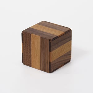 May include: A small wooden cube box featuring a geometric pattern of alternating light tan and dark brown wood grain panels. The box has a flat top and square sides, constructed from multiple vertical and horizontal strips of wood.