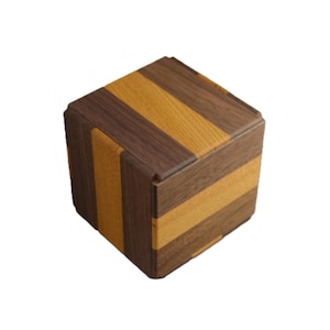 Puede incluir: Un cubo de madera con rayas alternas de madera marrón oscuro y marrón claro. El rompecabezas tiene una forma geométrica con una base cuadrada y una tapa. La veta de la madera es visible, agregando textura al diseño.