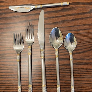 May include: A stainless steel flatware set with gold-colored accents. The set includes a knife, forks, and spoons. The handles have a textured design with gold bands at the ends. The knife is at the top of the image.
