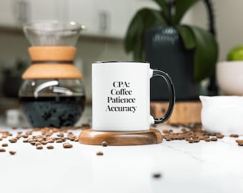 May include: A white coffee mug with a black handle and rim, featuring the text "CPA: Coffee, Patience, Accuracy." The mug is on a wooden coaster, with coffee beans scattered around. A glass coffee maker and a white creamer jug are also in view.