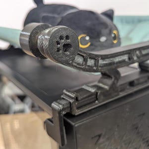 May include: A black cat-shaped plush toy rests on a black, 3D-printed cat bed. The bed has a curved design with a paw print detail on the end. A light blue support bar extends from the bed.