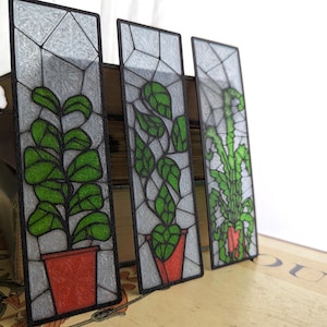 May include: Three rectangular stained glass bookmarks featuring green plants in red pots. The glass has a geometric pattern. The bookmarks are displayed on a stack of books, suggesting a botanical or literary theme.