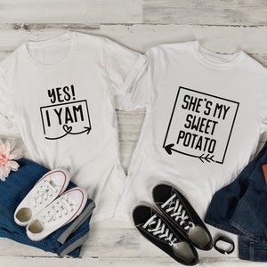 May include: Two white t-shirts with a couple's matching design. The first shirt says "YES! I YAM" with a heart and arrow. The second shirt says "SHE'S MY SWEET POTATO" with a heart and arrow.