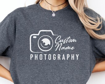 May include: Heather dark grey t-shirt with a white camera graphic and the words "Custom Name Photography". The shirt is made of a soft material and is a casual style.