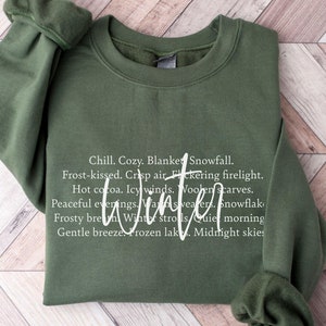 May include: A green sweatshirt with a white graphic print that reads "Chill. Cozy. Blanket Snowfall. Frost-kissed. Crisp air. Flickering firelight. Hot cocoa. Icy winds. Woolen scarves. Peaceful evenings. Winter strolls. Quiet morning. Gentle breeze. Frozen lake. Midnight skies."