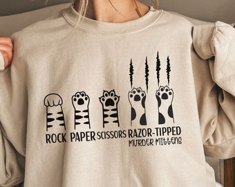 May include: Beige sweatshirt featuring a black graphic of cat paws representing rock, paper, scissors, and razor-tipped murder mittens. The text "ROCK PAPER SCISSORS RAZOR-TIPPED MURDER MITTENS" is printed below the paws. The sweatshirt has long sleeves.