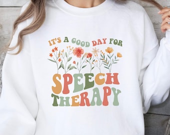 May include: White sweatshirt with the text "It's a good day for Speech Therapy" in colorful, retro-style lettering. Floral accents are above the text. The sweatshirt is a long-sleeved, crew neck style.