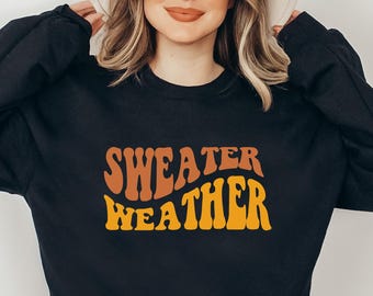 May include: Black sweatshirt with the text "SWEATER WEATHER" in orange and brown retro font. The sweatshirt is a crew neck style. The model is wearing a cream-colored hat.