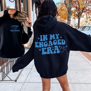 May include: A black hoodie with a blue graphic that says "In My Engaged Era" with a ring and hearts. The hoodie has a drawstring hood and a kangaroo pocket.