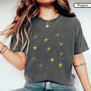 May include: A charcoal gray t-shirt featuring a pattern of yellow four-pointed stars and sparkles across the chest. The shirt has a crew neckline and short sleeves. The word Pepper is printed in a small white box in the top right corner.