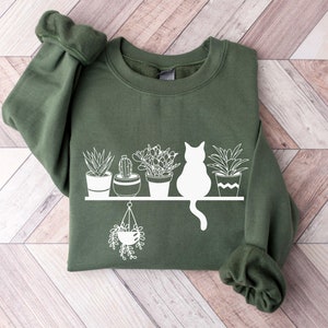 May include: A dark green sweatshirt with a white graphic of a cat sitting on a shelf with potted plants. The plants include a cactus, a succulent, and a hanging plant.