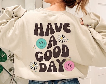 May include: A cream-colored sweatshirt with the text "HAVE A GOOD DAY" in brown letters with a retro font. The text is surrounded by colorful smiley faces and flowers.