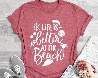 May include: Heather pink t-shirt with the text "Life is better at the beach!" in white. The shirt features a variety of beach-themed icons, including a seashell, a palm tree, a beach umbrella, sunglasses, and a dolphin.