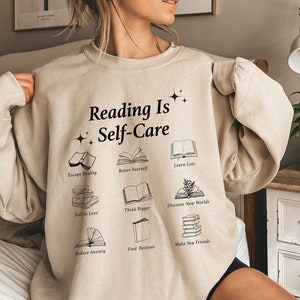 May include: A beige sweatshirt with the text "Reading Is Self-Care" and illustrations of books with phrases like "Escape Reality", "Better Yourself", "Learn Lots", "Fall in Love", "Think Bigger", "Discover New Worlds", "Reduce Anxiety", "Find Passions", and "Make New Friends".