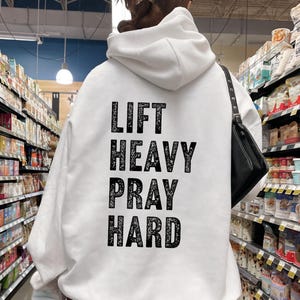 May include: White hoodie with a black print that says "Lift Heavy Pray Hard". The person is wearing the hoodie in a grocery store.