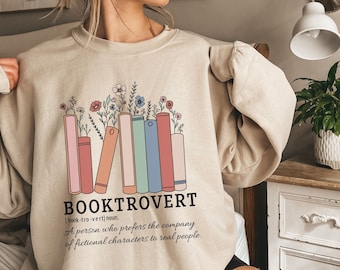 May include: A beige sweatshirt with a graphic design of a stack of books with flowers. The text "BOOKTROVERT" is written in black with a definition below: "[book-tro-vert] noun. A person who prefers the company of fictional characters to real people."