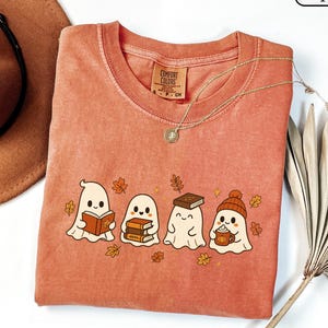 May include: A yam-colored crewneck t-shirt featuring a graphic of four white ghosts. One ghost reads a book, one holds a stack of books, one has a book on its head, and one wears a beanie while holding a mug. Scattered orange autumn leaves surround the ghosts.
