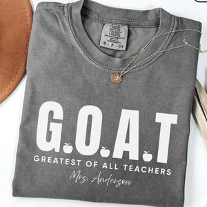 May include: A charcoal gray t-shirt with white text that reads G.O.A.T. GREATEST OF ALL TEACHERS and Mrs. Anderson. The design includes three small white apple icons between the letters. The shirt has a crew neckline and a Comfort Colors label.