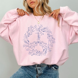 May include: A pink sweatshirt with a blue peace sign design surrounded by floral details. The peace sign is made of two hands with flowers and leaves growing around them.