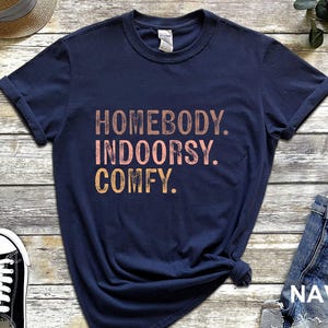 以下が含まれることがあります： ネイビーブルーのTシャツに「Homebody. Indoorsy. Comfy.」の文字がピンクとオレンジでプリントされています。