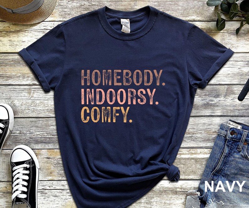 以下が含まれることがあります： ネイビーブルーのTシャツに「Homebody. Indoorsy. Comfy.」の文字がピンクとオレンジでプリントされています。