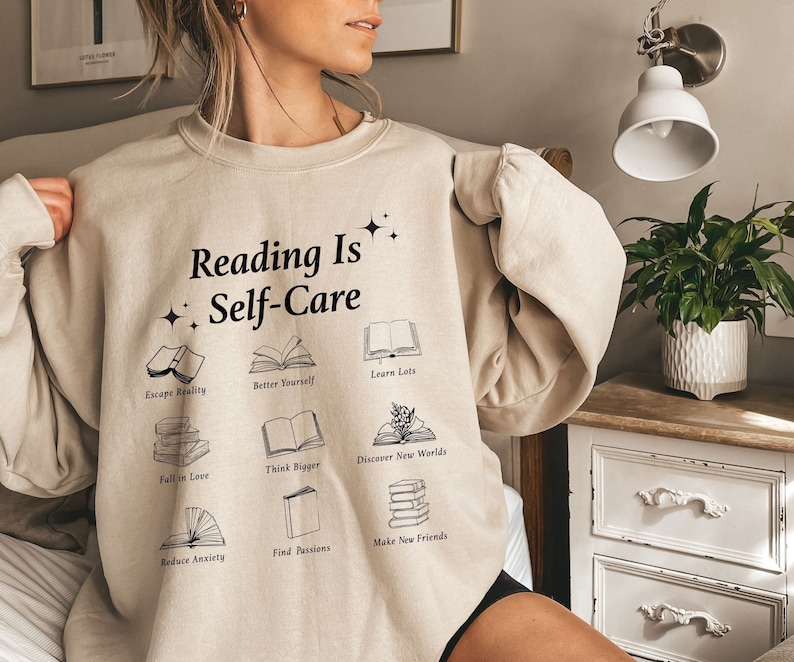 May include: A beige sweatshirt with the text "Reading Is Self-Care" and illustrations of books with phrases like "Escape Reality", "Better Yourself", "Learn Lots", "Fall in Love", "Think Bigger", "Discover New Worlds", "Reduce Anxiety", "Find Passions", and "Make New Friends".