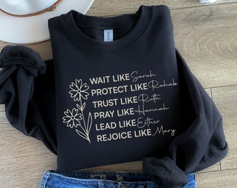 Op de afbeelding: Zwart sweatshirt met een gouden print van een bloemenmotief en de tekst "WAIT LIKE Sarah PROTECT LIKE Rahab TRUST LIKE Ruth PRAY LIKE Hannah LEAD LIKE Esther REJOICE LIKE Mary"