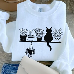May include: White sweatshirt with a black graphic of a cat sitting on a shelf with five potted plants. The plants are a cactus, a succulent, a fern, and two other plants. The cat is looking to the right.
