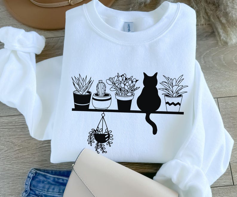 May include: White sweatshirt with a black graphic of a cat sitting on a shelf with five potted plants. The plants are a cactus, a succulent, a fern, and two other plants. The cat is looking to the right.