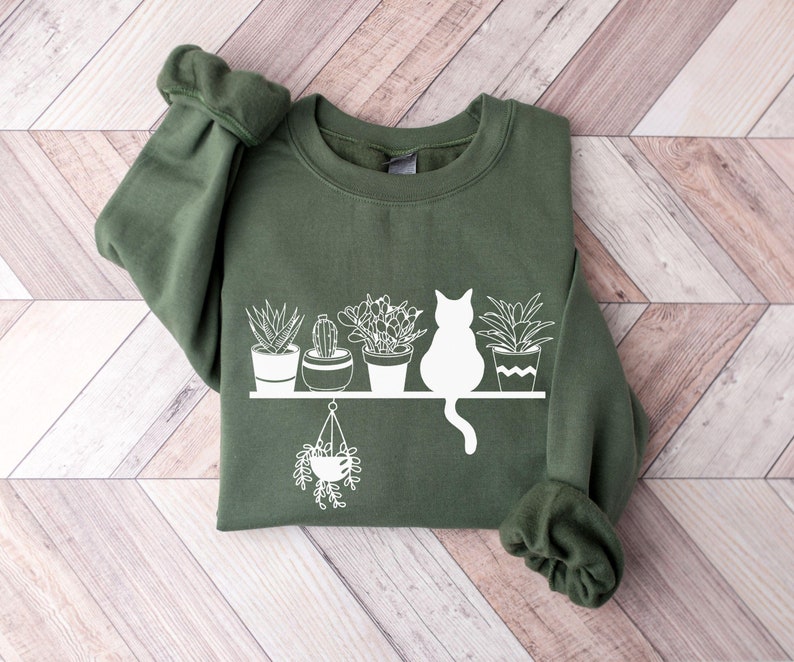 May include: A dark green sweatshirt with a white graphic of a cat sitting on a shelf with potted plants. The plants include a cactus, a succulent, and a hanging plant.